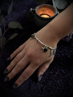 A silver-toned charm bracelet with a heart and angel motif worn on a person's wrist, set against a dark glittery background with a lit candle and leaves.