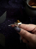 A hand with dark purple nail polish holds a silver-toned ring featuring a small golden-brown pyramid stone set in a silver bezel, against a dark, sparkly purple background with a cauldron prop.
