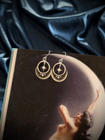 Silver earrings with star and moon designs on a dark surface with a woman reaching towards the moon.