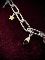 Silver chain bracelet with three charms, an arrow, a star, and a geometric point, on a dark fabric background.