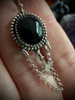 A close-up of a hand holding a silver-toned pendant necklace featuring a large oval black stone and dangling star-shaped chain charms.