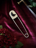 Giant stainless steel safety pin with a black crescent moon gemstone on a dark burgundy fabric background with flowers and a sword.
