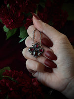Hand holding an ornate necklace with red gemstones against a dark floral background.