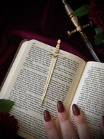 Open book with a sword-shaped bookmark and a hand holding it, on a dark background surrounded by flowers and a sword.