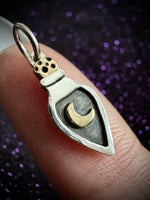 A close-up of a silver pendant shaped like a small potion bottle with a gold crescent moon and gold cap, held on a person's finger against a dark purple glittery background.