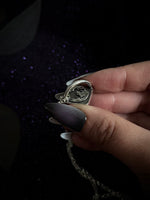 A close-up of a hand holding an open silver pendant with a chain, set against a dark background with subtle purple sparkles.