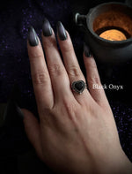 A hand with long black manicured nails wearing a gothic heart-shaped ring with a black onyx center and silver-toned border, set against a dark purple glittery background with a lit candle.