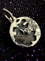 A close-up of a silver-toned round pendant charm with an embossed abstract design and a small loop at the top, photographed against a dark glittery purple background.