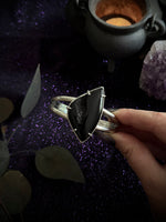 A hand holding a silver-toned cuff ring with a large, polished black triangular stone, set against a dark, mystical background with purple glitter, a cauldron, and crystals.