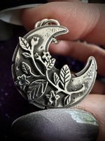 A close-up of a hand holding a crescent moon pendant with embossed floral and leaf details, shown against a dark purple sparkly background.