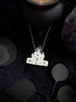 A silver pendant necklace featuring a house-shaped charm decorated with small stars and occult symbols, hanging from a silver chain. The necklace is styled on a dark glittery surface with dark leaves, a black ceramic bowl, and a purple crystal cluster.