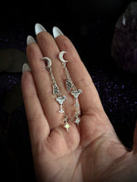 A pair of silver-toned dangle earrings with crescent moon, cocktail glass, and star charms, held in a person's hand against a dark glittery background.