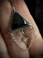 A close-up of a hand holding a silver-toned triangular pendant with a glossy black center and dangling star charms on a chain.