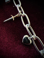 Silver chain bracelet with two charms, a dagger and a heart, on a dark fabric background.