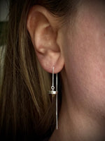 A close-up of a person's ear wearing a long, delicate silver dangle earring with a small circular charm and thin chain.