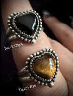 Two heart-shaped silver rings with textured beaded borders are shown worn on a hand. The top ring features a smooth black onyx stone, and the bottom ring features a golden-brown tiger's eye stone.