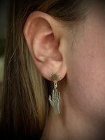 A close-up of a person's ear wearing a dangling earring with a small gold sunburst stud and a silver-toned hand-shaped charm.