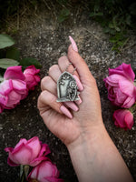 Hand holding a silver necklace with a gravestone pendant among pink flowers on a stone background