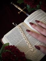 Open book with a decorative celestial bookmark and a hand holding it, surrounded by flowers and a sword on a dark background.
