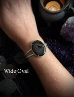 A close-up of a person's wrist wearing a silver-toned wide oval cuff bracelet with a dark agate stone centerpiece. The text 'Wide Oval' is overlaid on the lower left, with a dark mystical background featuring a candle, amethyst crystal, and dark leaves.
