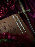 Silver and brass sword earrings on a leather book with a sword and flowers in the background.