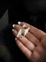 A close-up of a hand holding a pair of dangling earrings shaped like a wolf and a flame, with a small sun charm at the top, against a dark sparkly background.