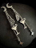 A pair of silver dangling earrings with crescent moon and cocktail-themed charms, displayed on a dark textured stone surface.