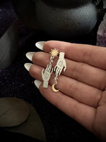 A pair of gold-toned dangle earrings held in a hand. One earring features a raised hand with a dangling sunburst, and the other features a raised hand with a dangling crescent moon.