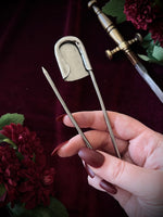 Hand holding the backside of a giant steel safety pin against a dark background with flowers and a sword.