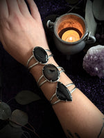Three silver cuff bracelets with large black stone inlays worn on a person's wrist, shown against a dark purple glittery background with a candle and crystals.
