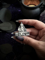A hand with long manicured nails holds a silver-toned metal charm shaped like a gothic haunted house, set against a dark, mystical background with a cauldron and purple glitter.