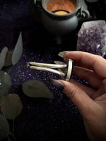 A hand with dark manicured nails holds a silver-toned metal hair claw clip against a dark, mystical background with a cauldron, purple glitter, and crystals.