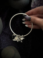 A silver bangle bracelet with charms, held by a hand with long dark and white manicured nails against a dark glittery background with decorative stones.