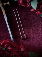 Silver chain earrings on a dark fabric background surrounded by a sword with red flowers.