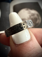 Silver ring with intricate design held by a hand on a white surface.
