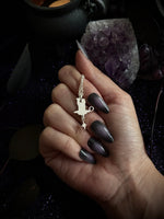 A hand with dark metallic nails holds a silver-toned keychain featuring a miniature cauldron and star charm, set against a dark mystical background with an amethyst cluster.