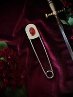 Giant stainless steel safety pin with a red gemstone on a dark burgundy fabric background with flowers and a sword.