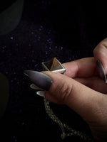 A close-up of a hand with long metallic silver nails holding a silver pyramid pendant necklace with a gold-toned center, set against a dark glittery background.
