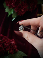Hand holding a silver necklace with a small dragon pendant against a dark background with red flowers.