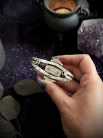 A hand holding a silver-toned metal bracelet with a star and crescent moon design, set against a dark background with purple glitter and mystical props.
