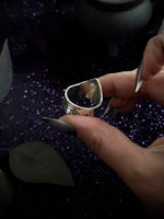 A close-up of a hand holding a silver-toned ring with a textured, hammered finish and small cutout details, set against a dark background with purple glitter.