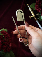 Hand holding the backside of a giant steel safety pin against a dark background with flowers and a sword.