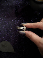 A close-up of a hand with long dark gray manicured nails holding a silver-toned ring with a textured, patterned band against a dark, sparkly purple background.