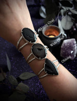 Three silver-toned cuff bracelets with black stone settings worn on a person's wrist, shown against a dark purple glittery background with decorative leaves, a candle, and a crystal. Text labels indicate Skinny Oval, Wide Oval, and Oblong.