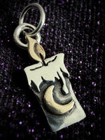 A silver wax candle pendant charm with a raised gold crescent moon and flame detail, shown on a dark glittery background.