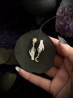 A hand holds a dark leaf displaying a pair of silver-toned hands earrings, one holding a sun and the other a crescent moon, against a dark, mystical background with crystals.