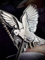 A close-up of a silver-toned dove pendant or brooch with wings spread, attached to a chain, held against a dark background.