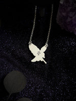 A silver bird pendant necklace with a small balance scale engraving, displayed on a dark glittery background with decorative leaves and crystals.