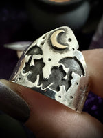 A close-up of a hand holding a wide silver ring with cutout witch motifs, including a crescent moon and stars, featuring a small gold crescent accent.
