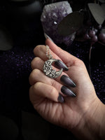 A hand with long dark gray manicured nails holds a silver crescent moon pendant with floral details and a matching chain. The background is dark with purple glitter and crystal elements.